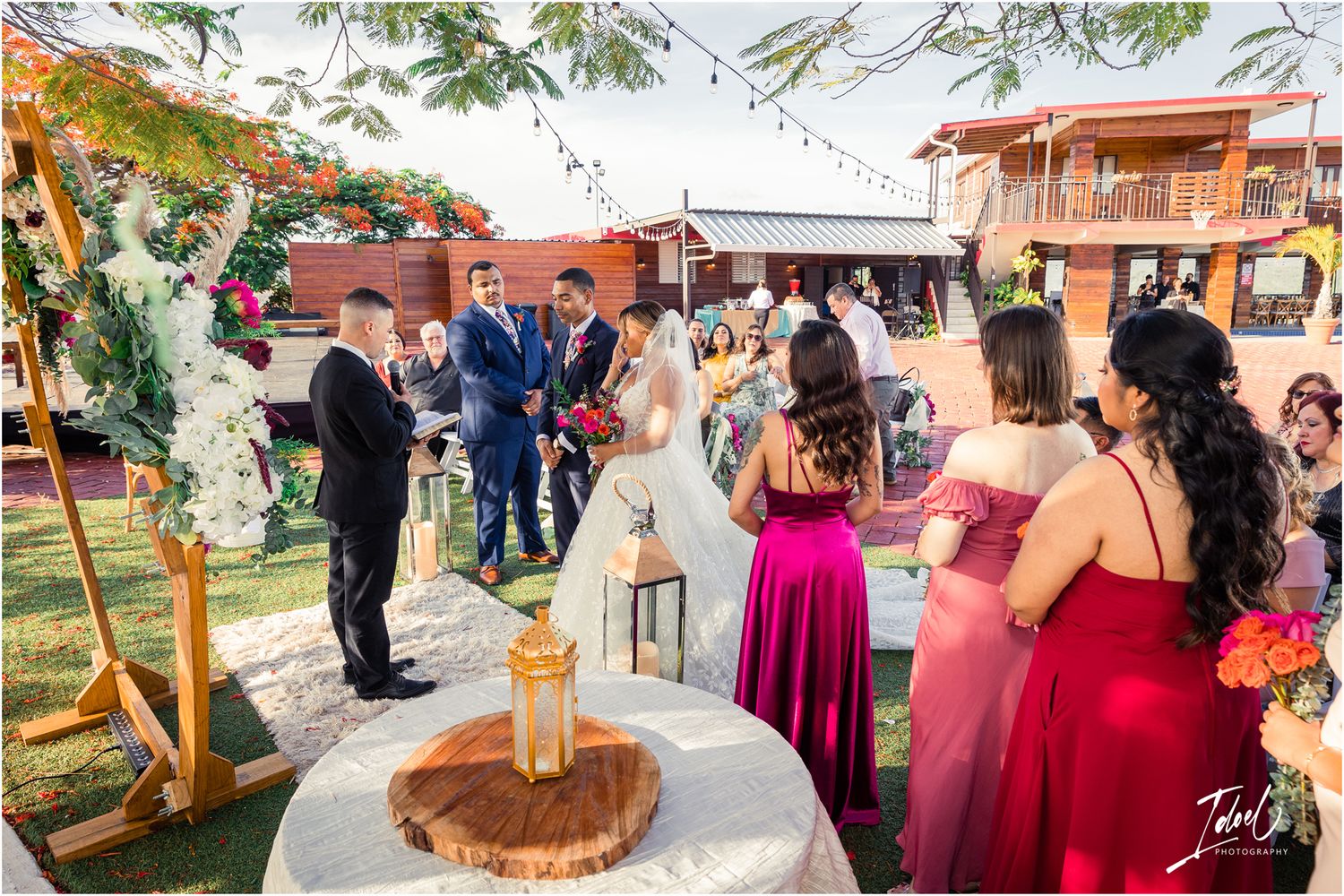 Outdoor wedding ceremony with burgundy bridesmaids dresses and rustic lantern decor on brick patio.