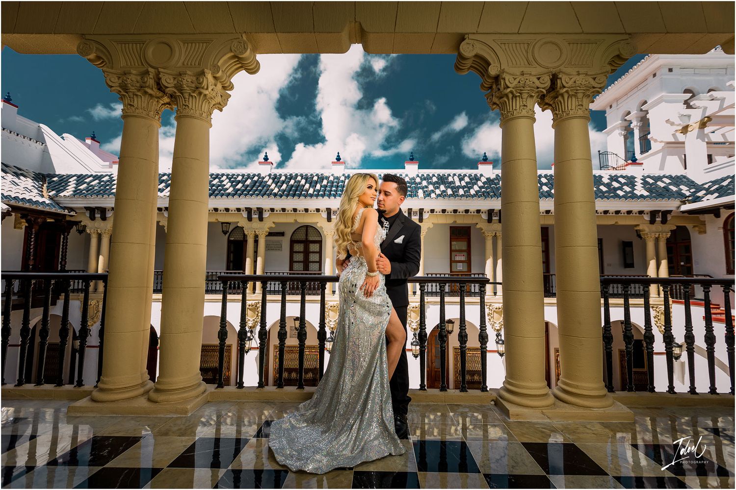 Elegant pose on balcony framed by ornate cream columns and blue sky.