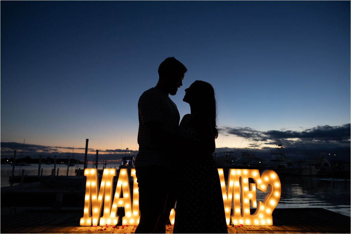 Silhouetted couple embraces in front of illuminated marquee letters at sunset against a darkening sky.