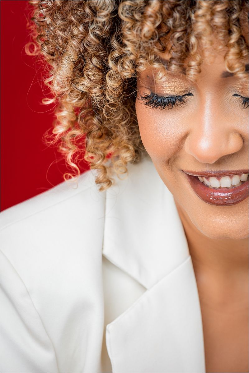 Close up portrait showing natural curly blonde hair and bright smile against red background wearing white blazer.