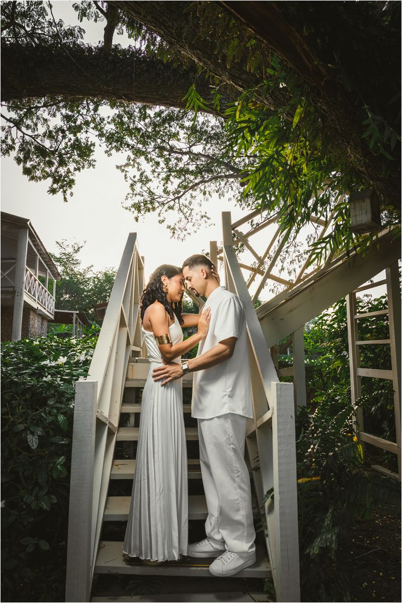 A romantic couple in light colored formal attire share intimate moments on outdoor wooden steps surrounded by greenery.