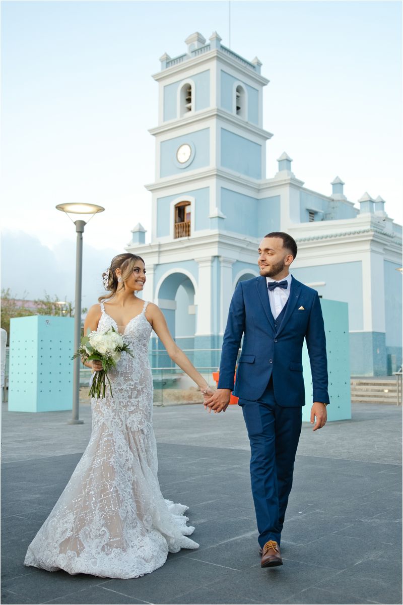 Bride and groom walk hand in hand before a blue church, she in lace gown, he in navy suit with bow tie.