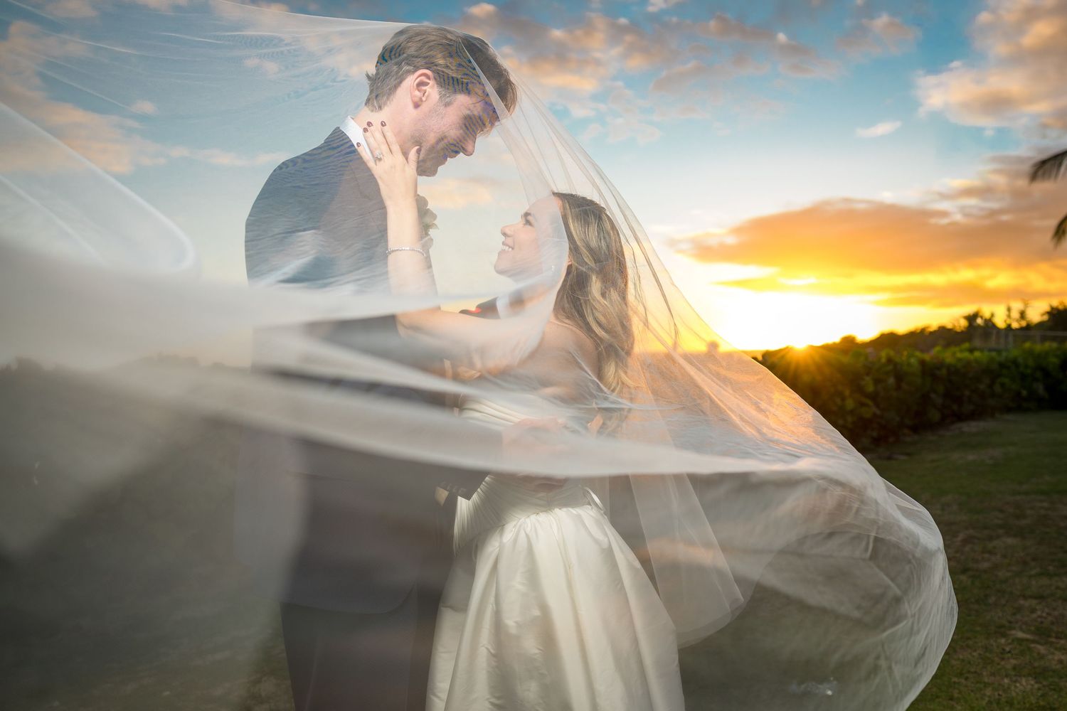 A romantic couple embraces in a flowing white dress during a golden sunset in an outdoor field.