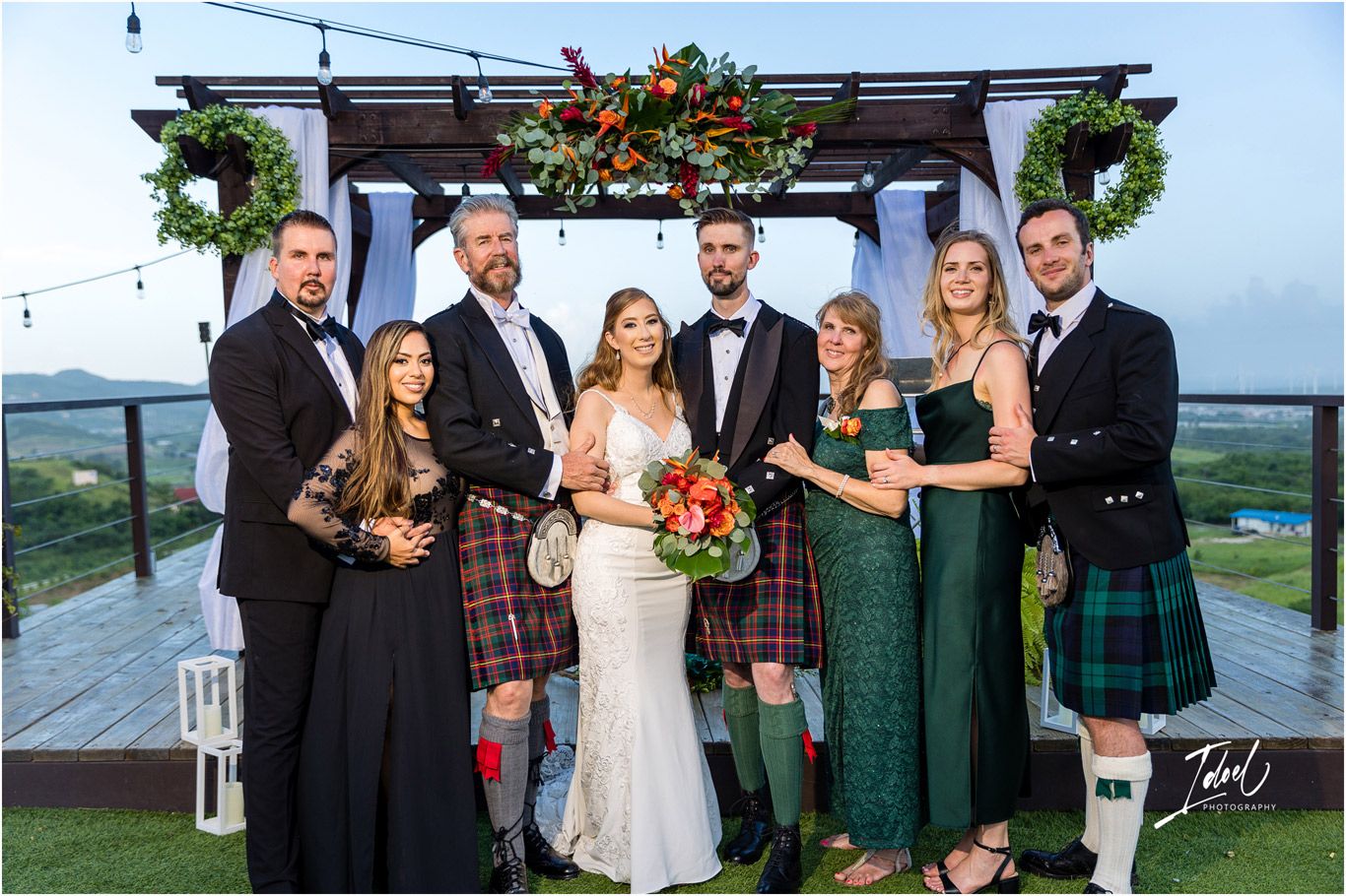 Wedding party in formal Scottish attire posing under an outdoor pergola with mountain views.