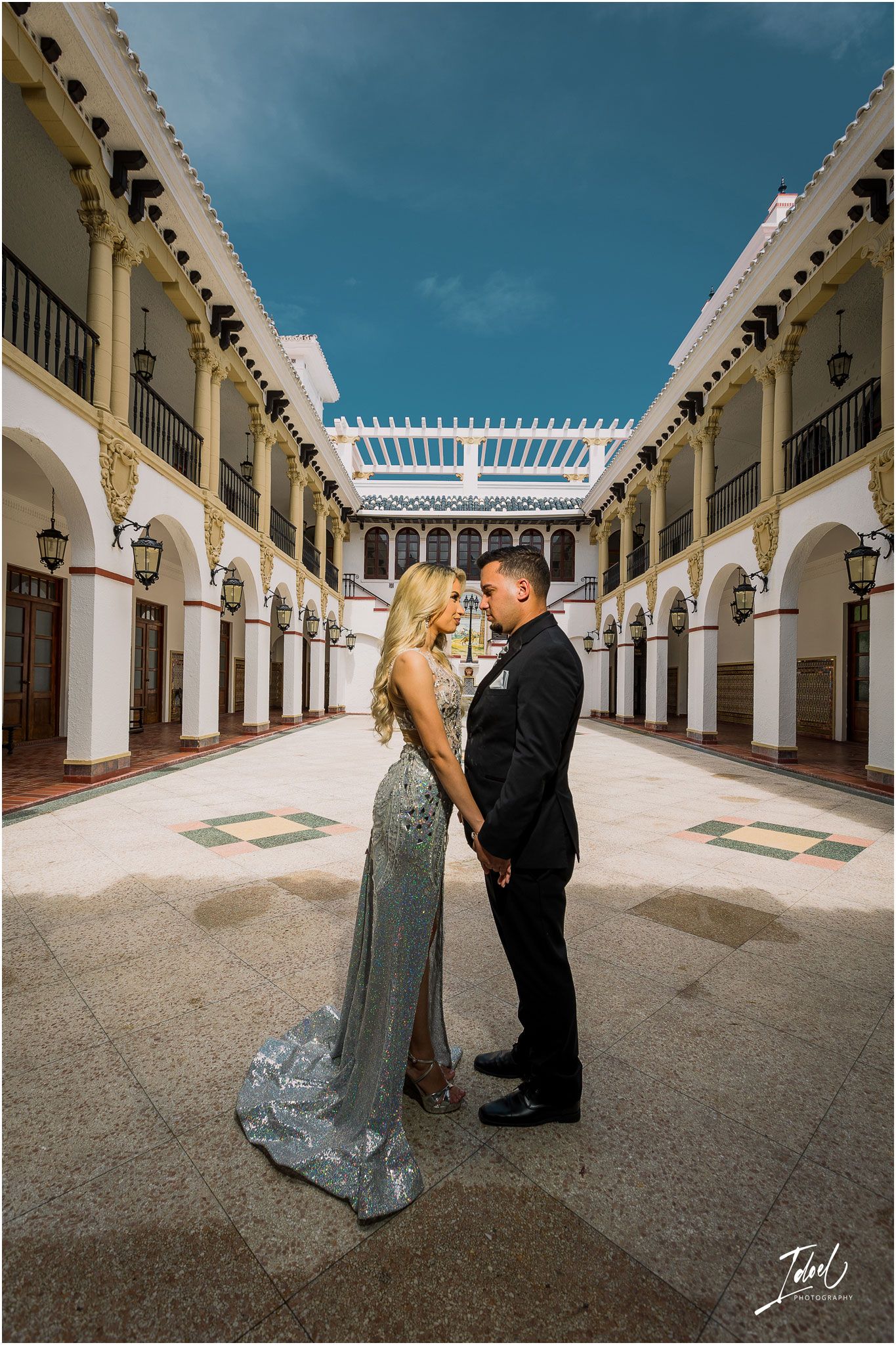 Couple posing in formal attire within a Spanish colonial courtyard featuring white columns and arches.