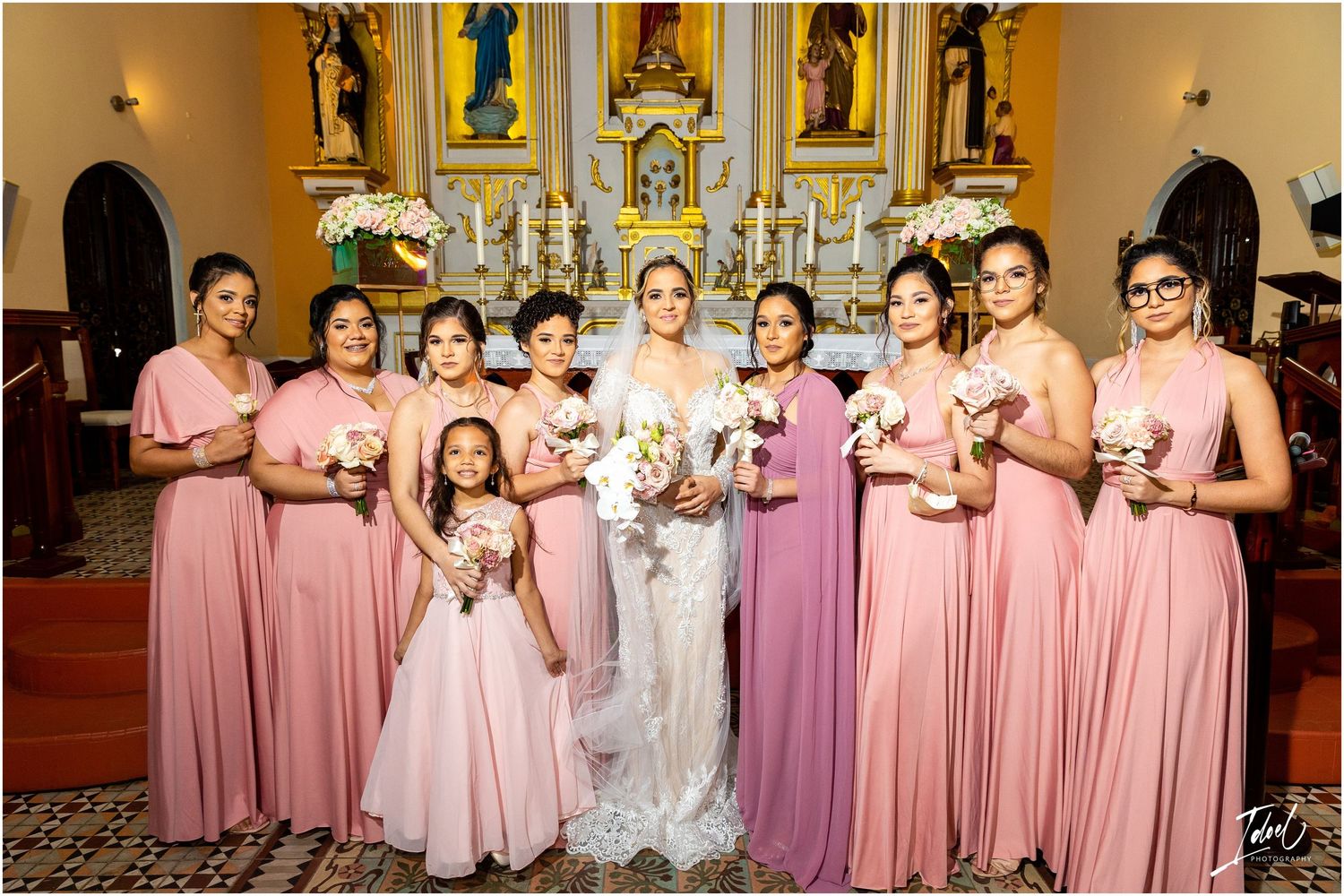 Wedding party wearing pink and purple bridesmaids dresses poses inside ornate church with golden altar.
