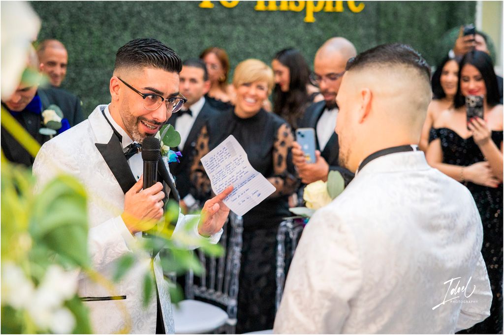 Two people exchange vows during an outdoor wedding ceremony as guests watch and take photos in the background.