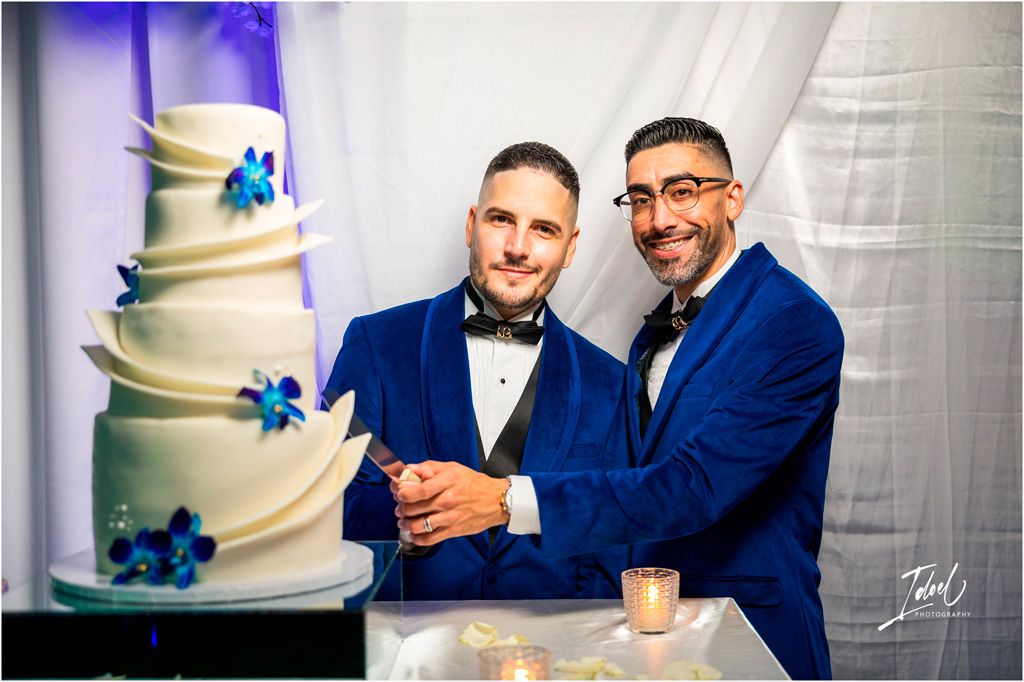 A beautiful multi-tiered white wedding cake decorated with blue flowers stands beside two people in matching navy blue suits.