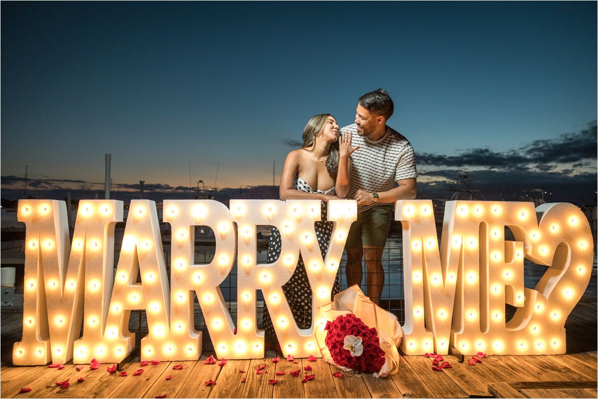 A romantic proposal scene features illuminated 'MARRY ME?' marquee letters on a wooden dock at dusk.