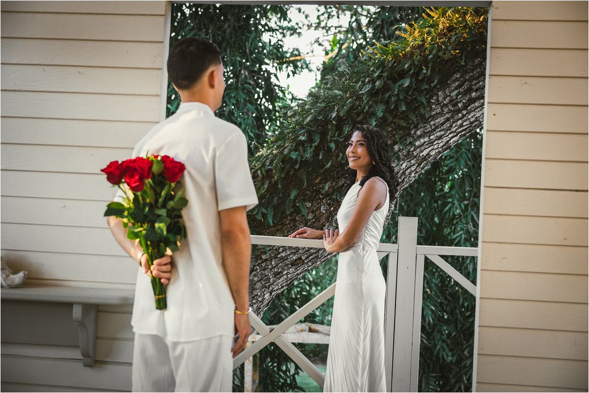 Couple in white attire standing together on a porch with floral decorations and greenery in the background.