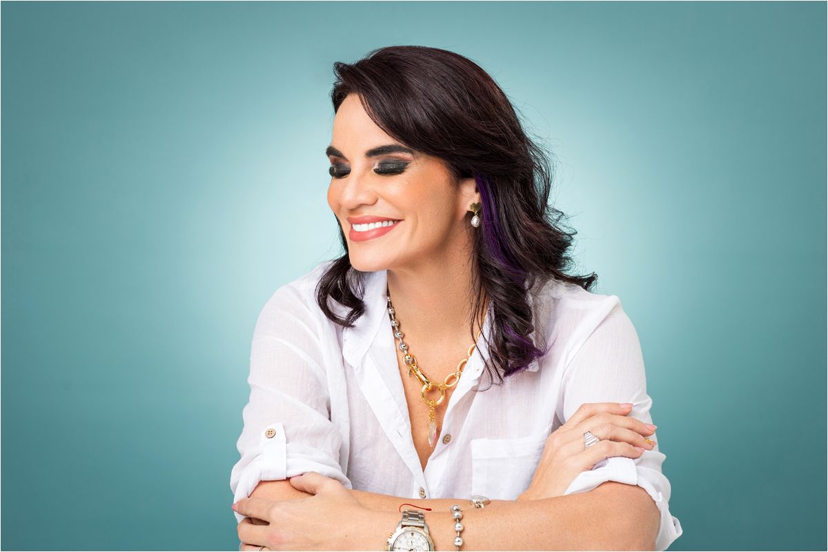A smiling woman with dark hair wearing a white button-down shirt and jewelry poses against a teal background.