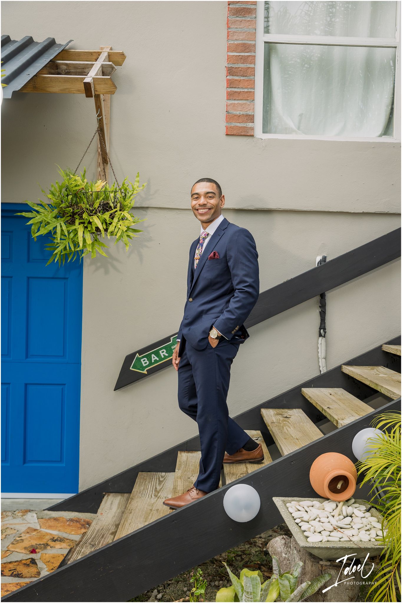 A person in a navy suit poses casually on wooden stairs next to a bright blue door and hanging plant.
