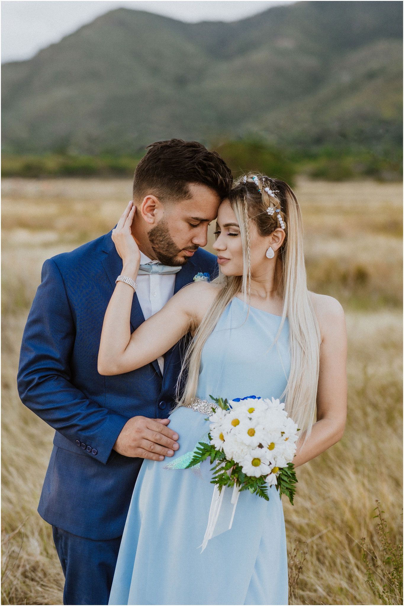 A romantic portrait of a couple in blue attire holding a daisy bouquet in a mountain field.