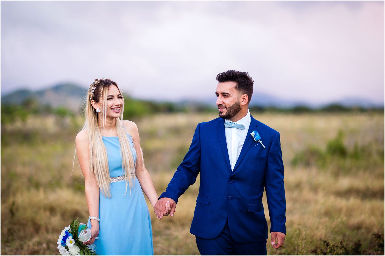 A joyful couple walks hand in hand through a grassy field with mountains in the distance.