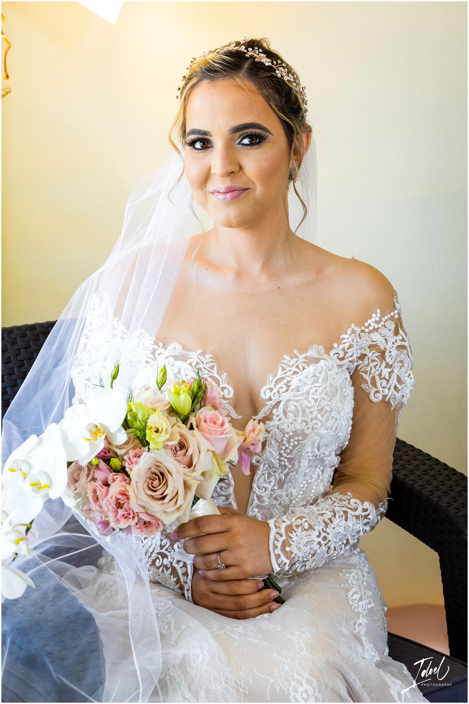 Bride wearing detailed lace gown holds pink and white bouquet while seated against neutral background.