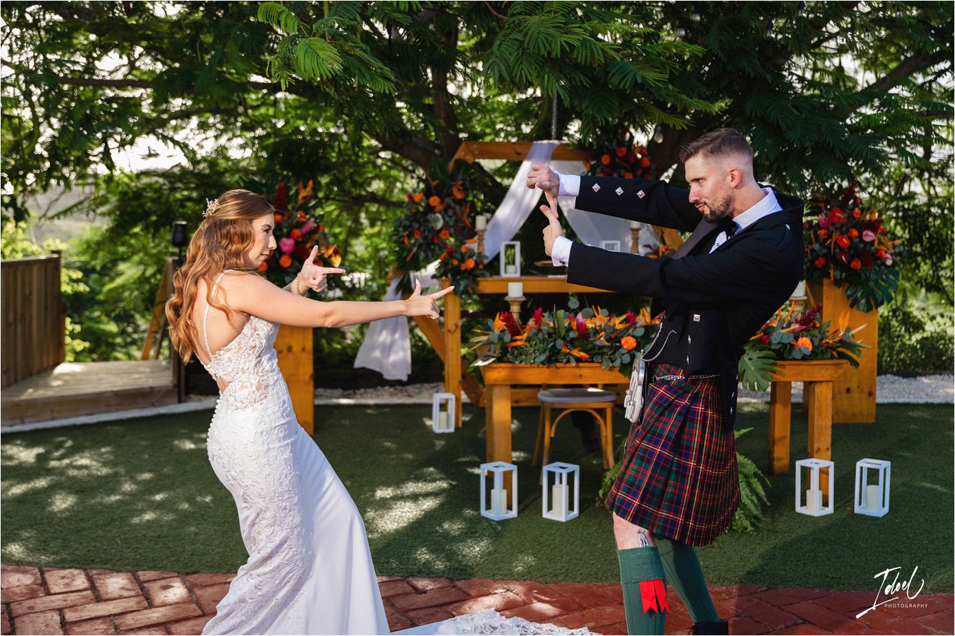 Joyful wedding couple dancing together in garden setting with decorated wooden arch in background.