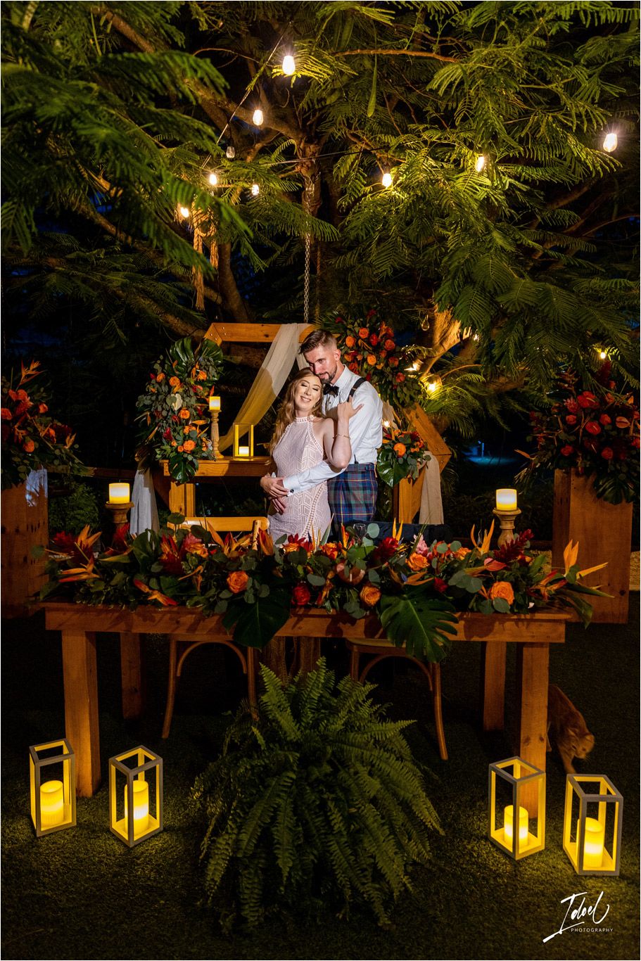 Evening reception setup with lanterns and floral arrangements under illuminated trees.