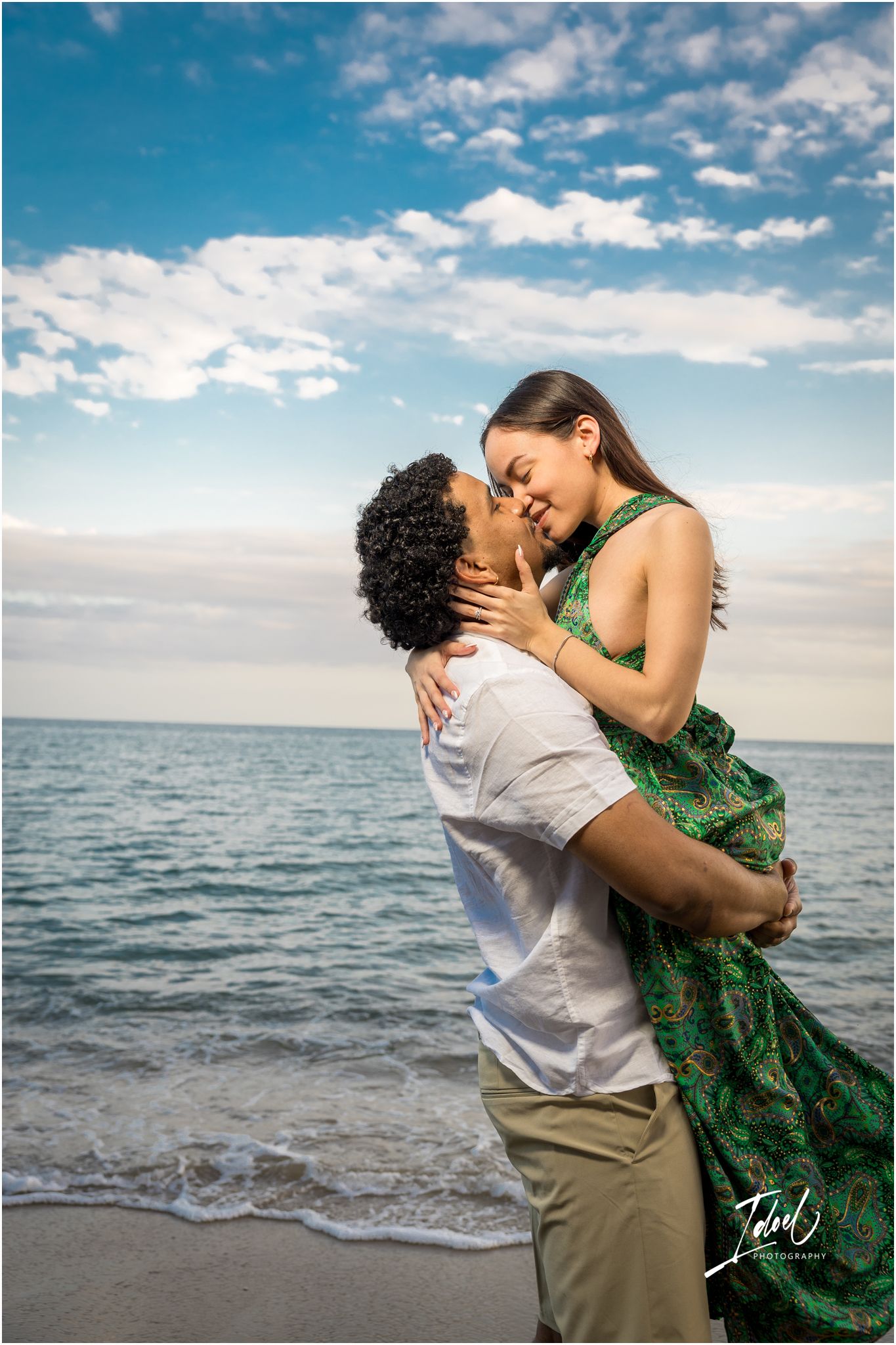 A romantic couple embraces on a beach with blue skies and ocean waves in the background.