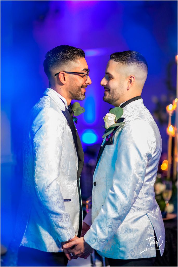 Two people in matching white tuxedo jackets share an intimate moment against a blue-lit backdrop during a celebration.