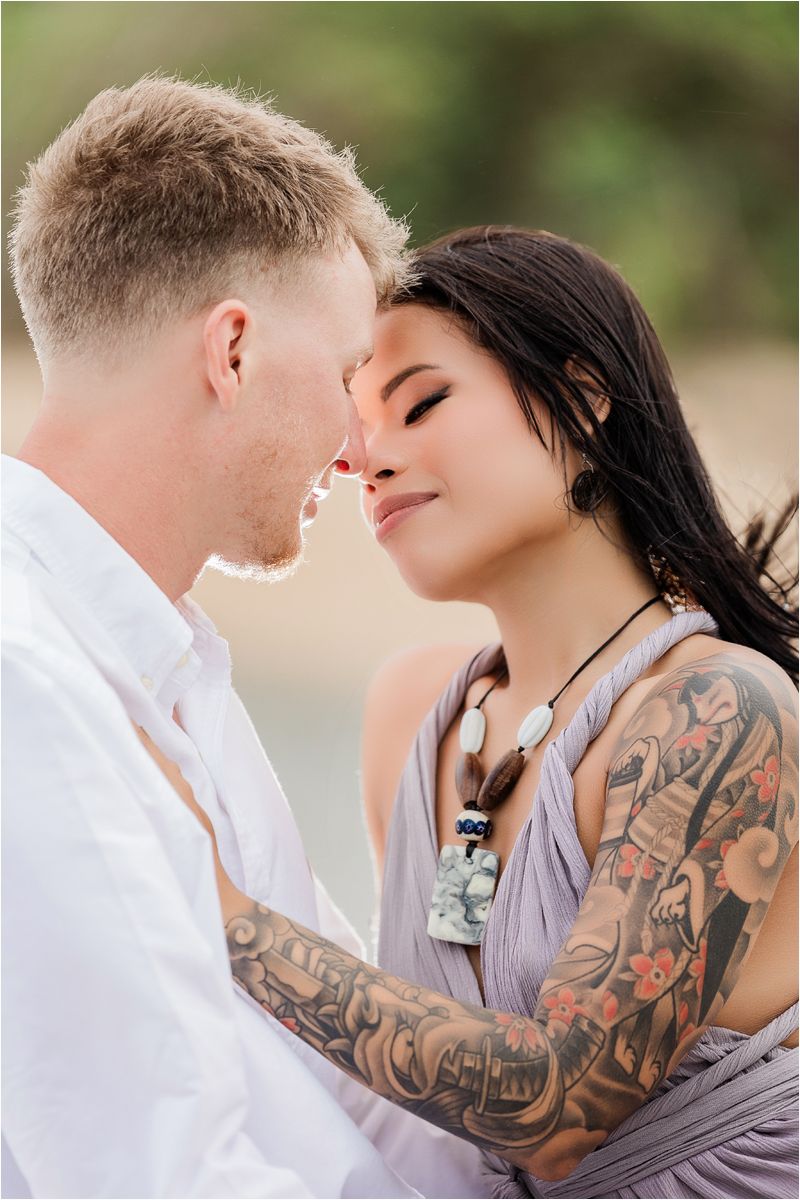 A romantic couple embraces on a beach, with the woman wearing a lavender dress and displaying intricate arm tattoos.