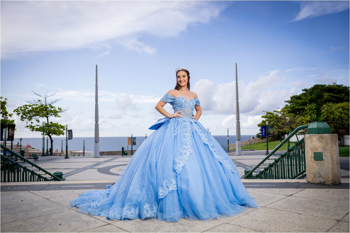 A person in a flowing light blue ball gown poses outdoors against a waterfront backdrop with ornate lampposts.