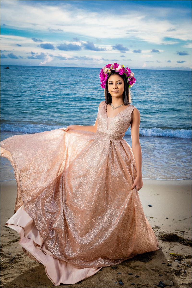 A person in a rose gold sequin gown poses gracefully on a beach at sunset with the ocean waves in the background.