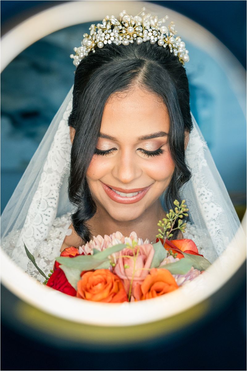 Beautiful bride wearing pearl crown and veil smiles while holding vibrant orange and pink rose bouquet.