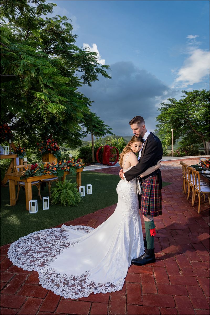 Wedding couple embracing on red brick patio with dramatic train of white lace gown.