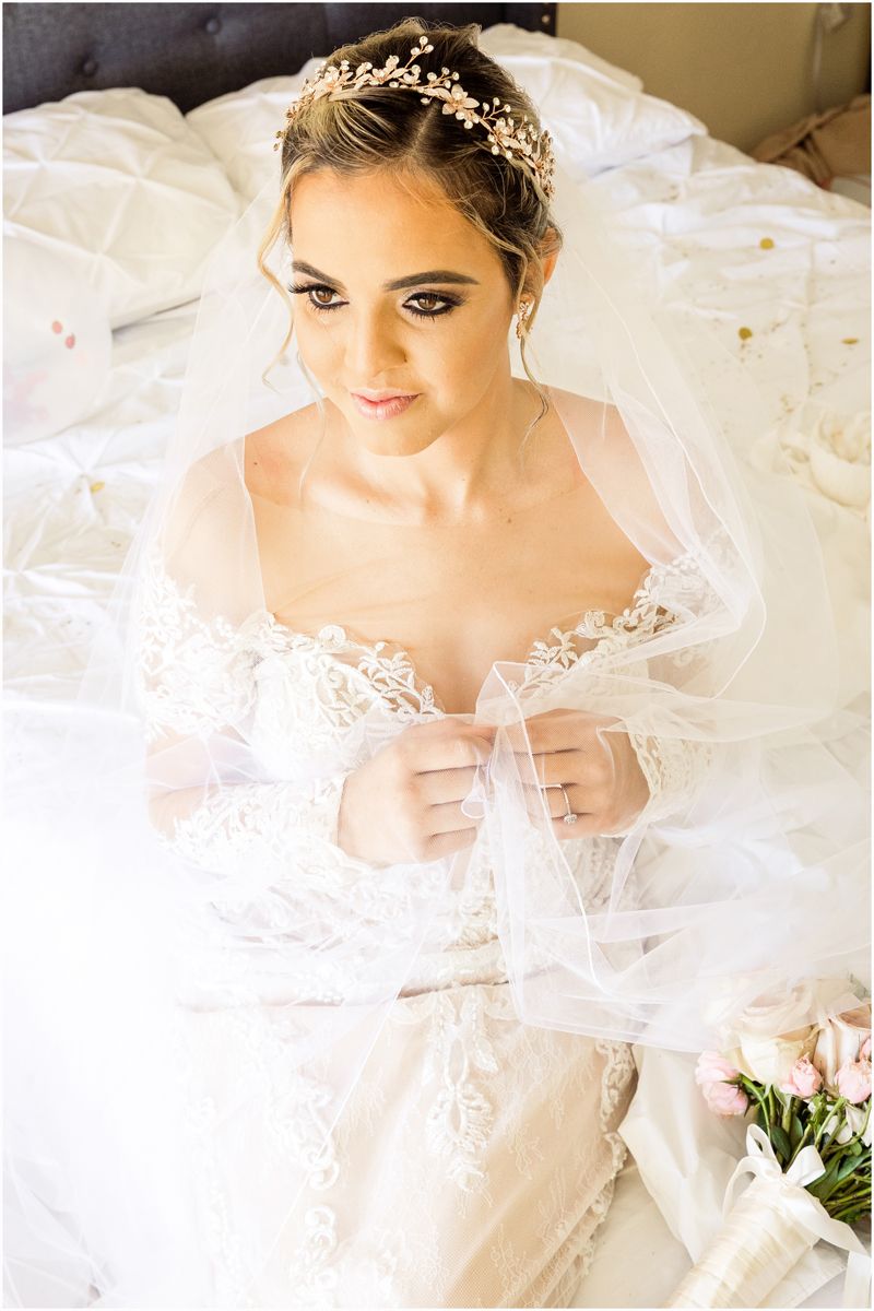 Bride in an elegant lace wedding dress with a crystal headpiece and veil sitting on white bedding.