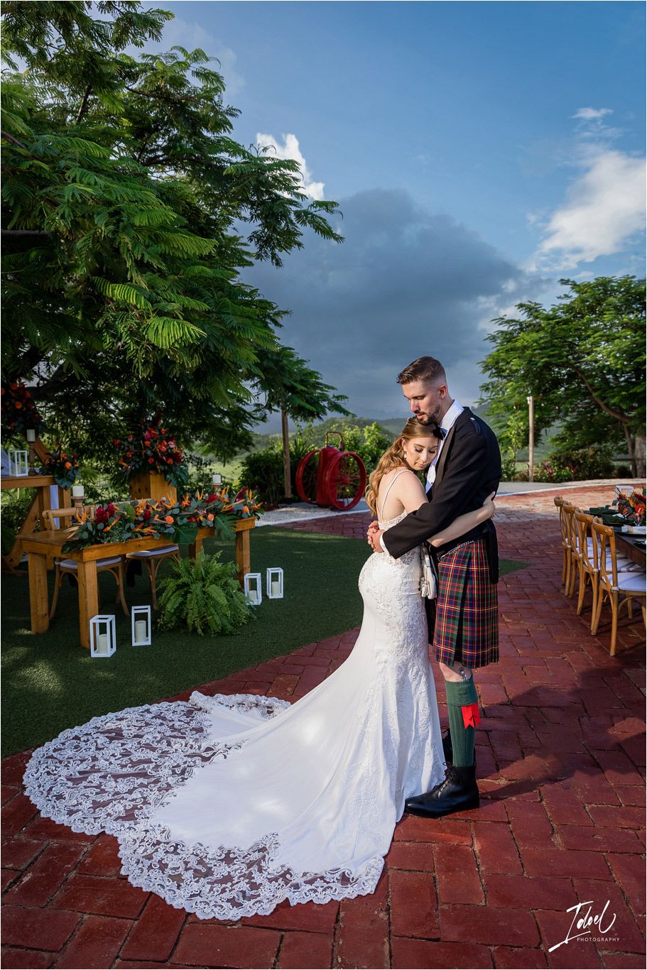 A Scottish wedding couple embraces on a brick patio surrounded by lush greenery and outdoor reception decor.