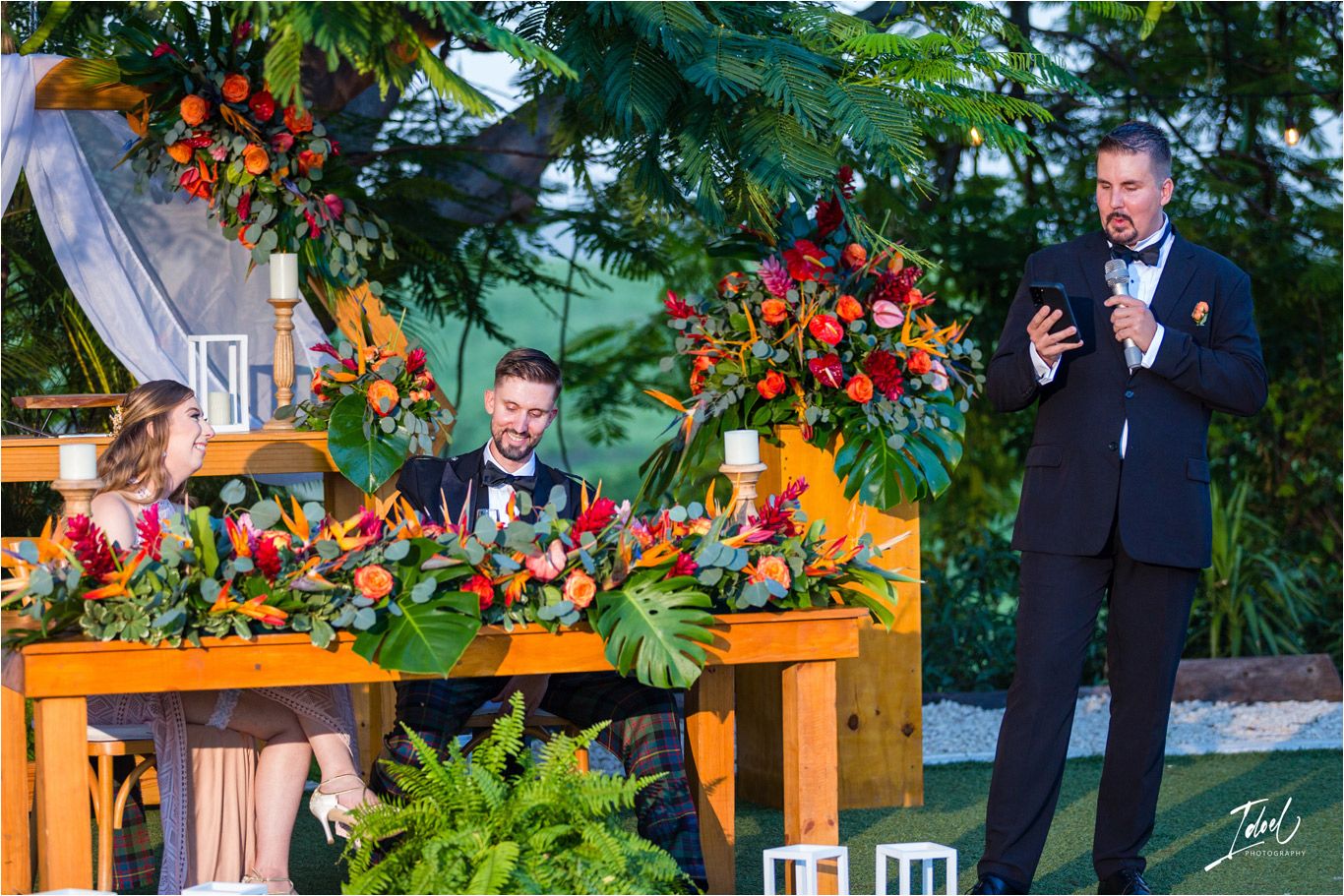 Outdoor wedding reception with rustic wooden tables decorated with tropical floral arrangements.