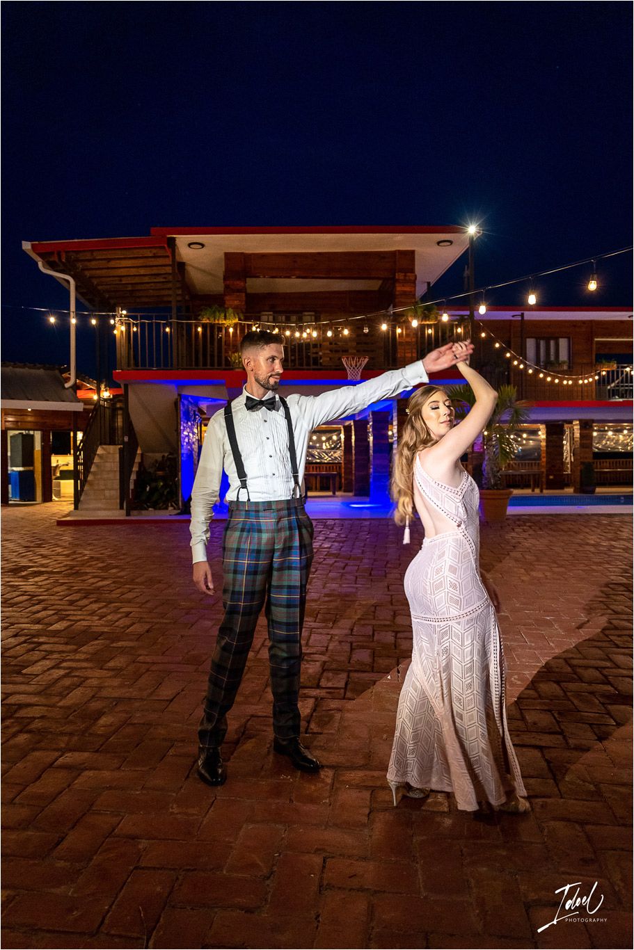 A couple dances together at night under string lights on an outdoor brick patio.