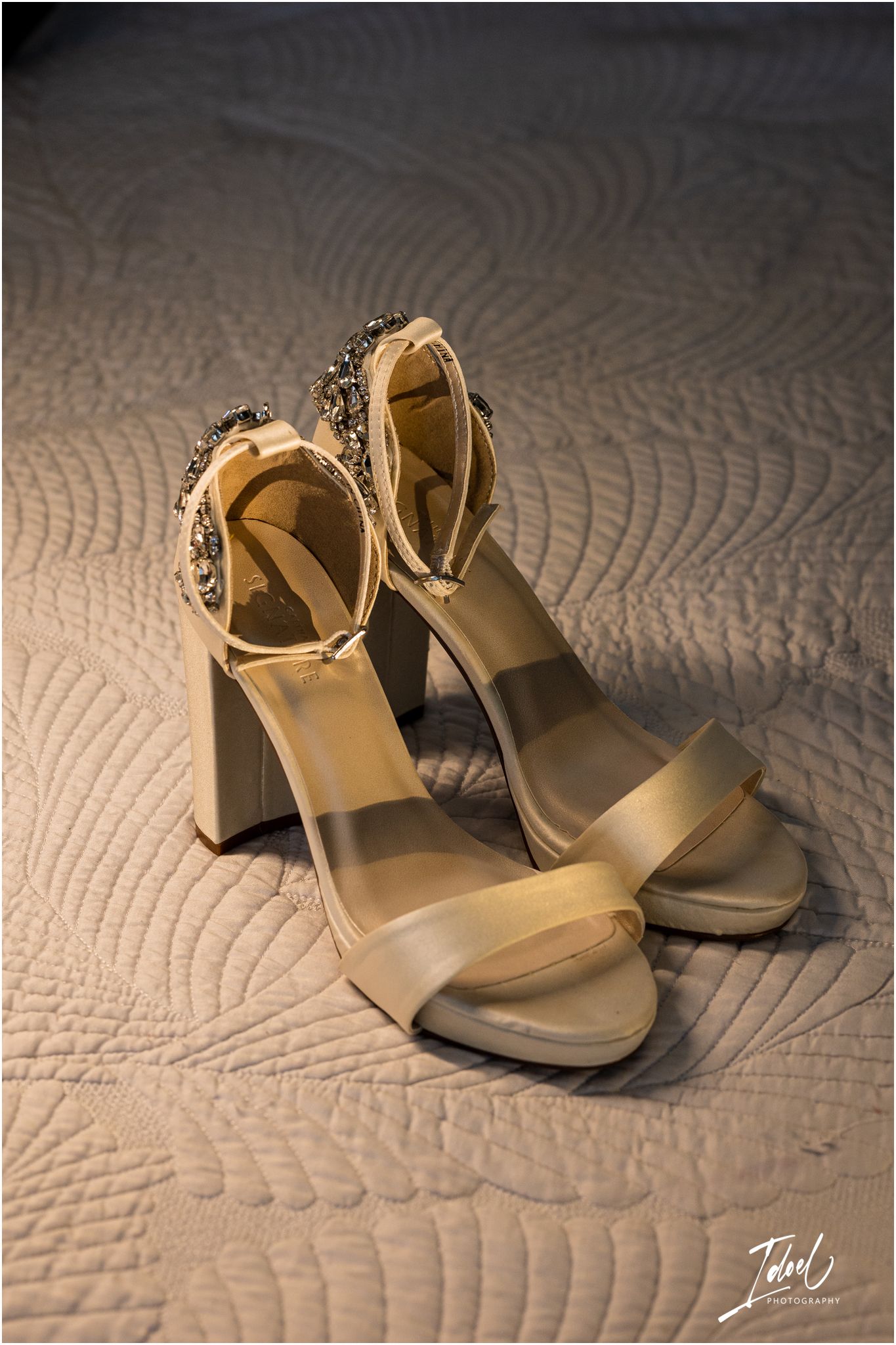 Elegant beige high heel sandals with crystal embellishments photographed on a quilted fabric background.