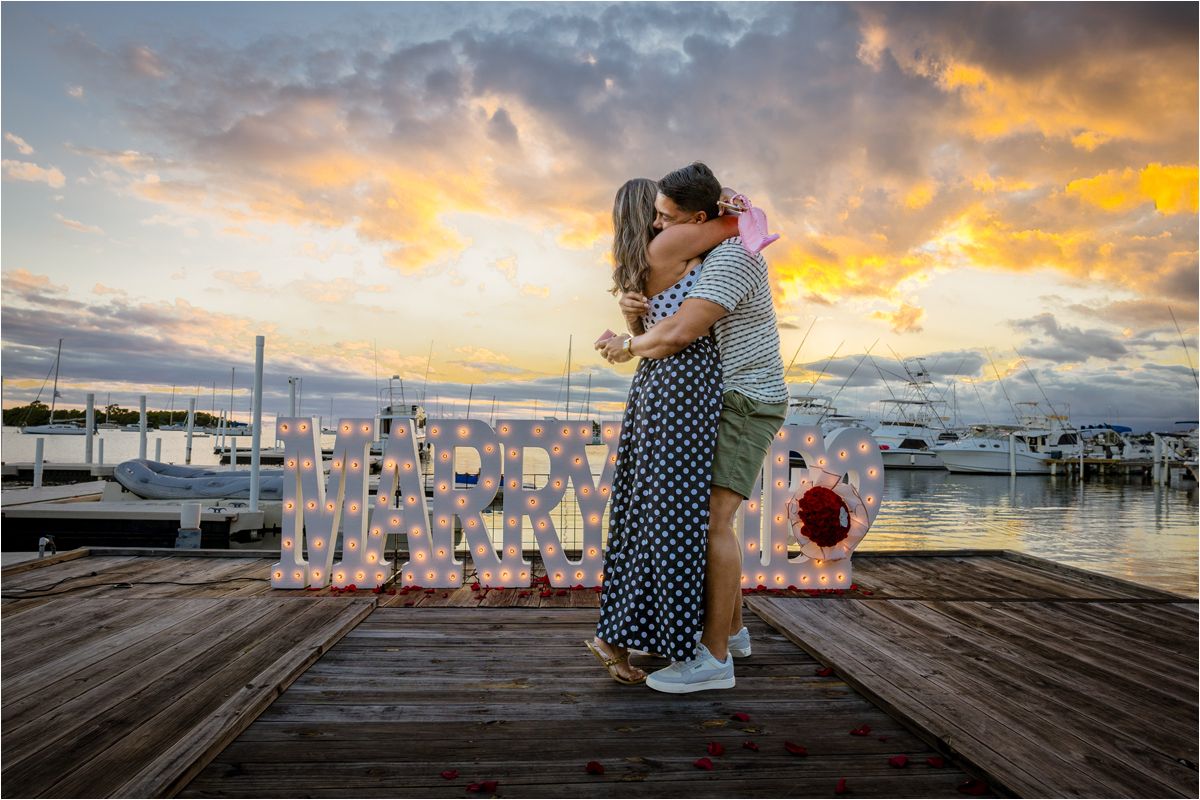 A romantic sunset proposal moment on a marina dock with glowing letter lights and boats in the background.
