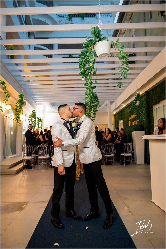 Two people in matching white tuxedo jackets share a romantic moment during their wedding ceremony on a navy blue aisle runner.