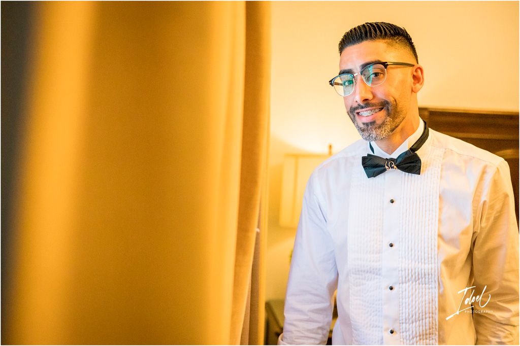 A groom in a crisp white tuxedo shirt and black bow tie smiles warmly before his wedding ceremony.