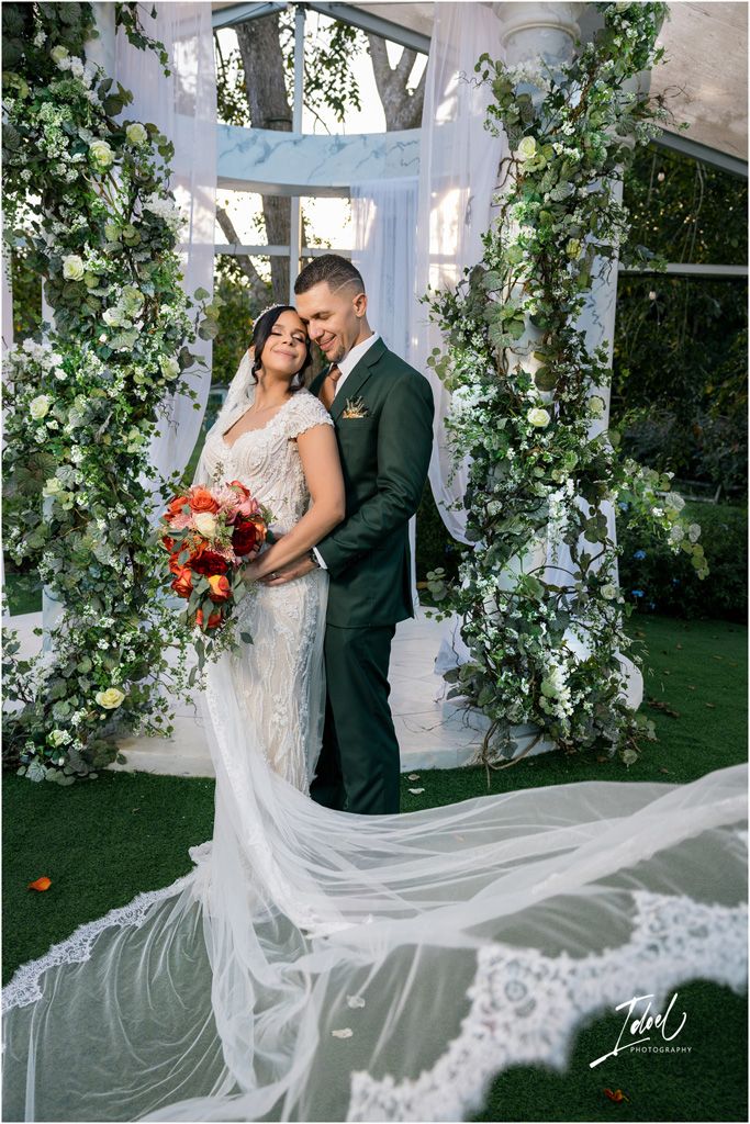 Couple sharing romantic moment at elegant outdoor wedding ceremony with floral arch and flowing lace veil.