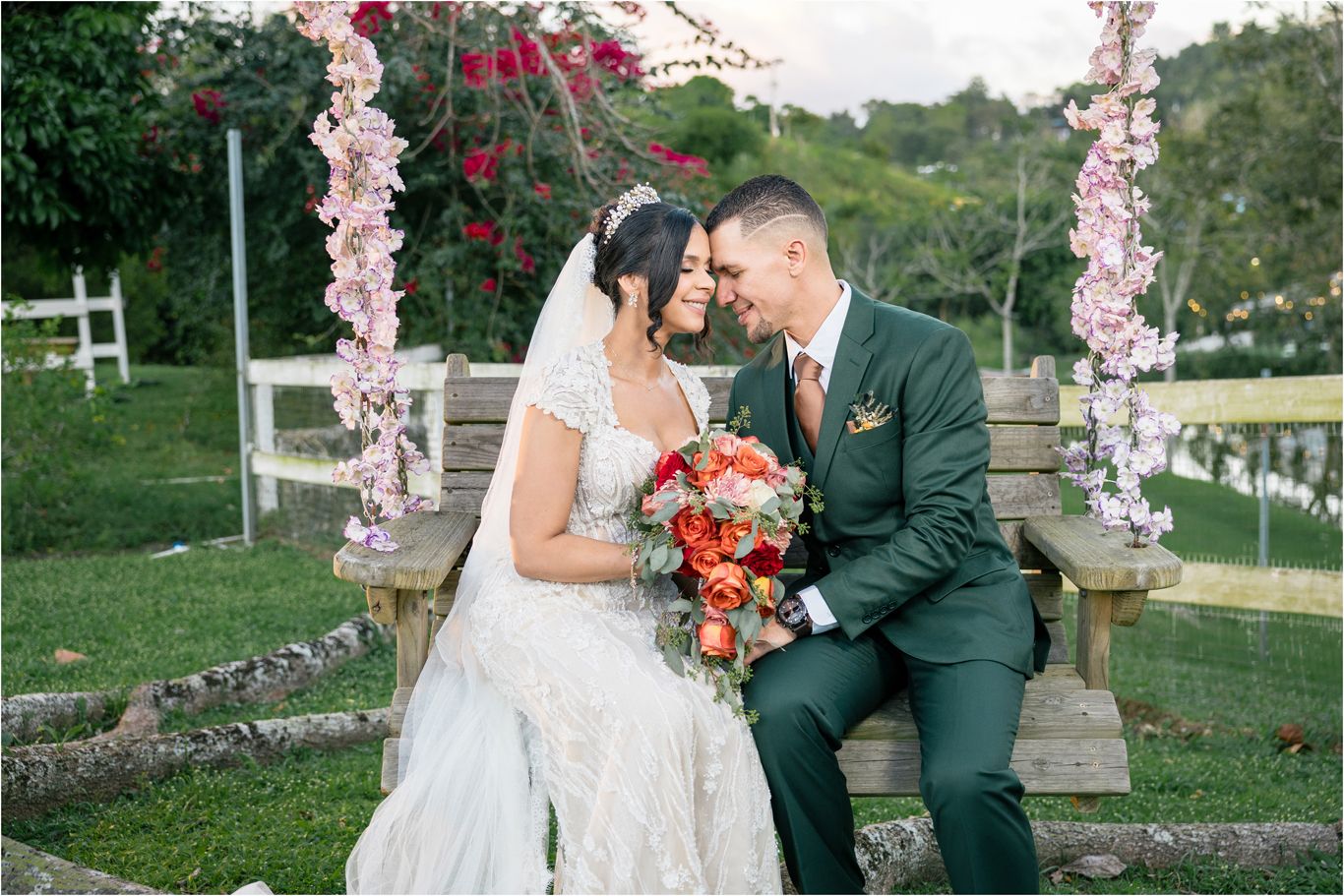 A couple shares a romantic moment on a rustic bench under a floral arch decorated with pink blossoms during an outdoor ceremony.