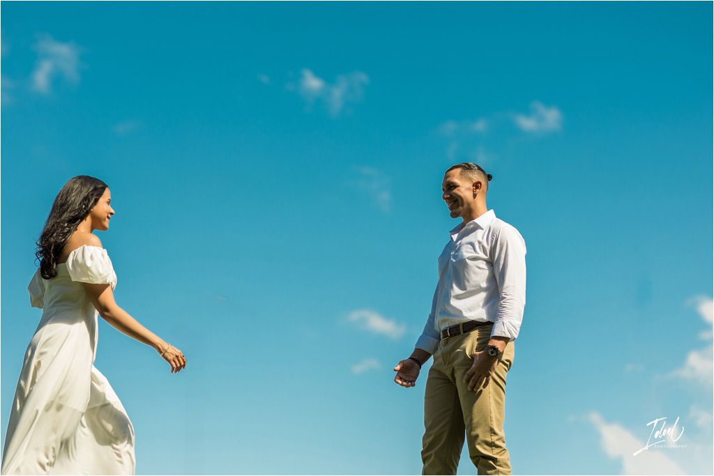 A couple in light colored outfits having a playful interaction against a bright blue sky backdrop.