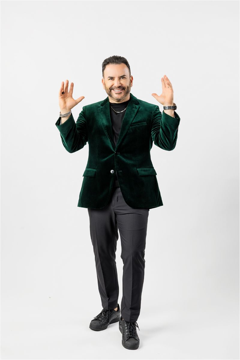A person in a green velvet blazer and black pants gestures enthusiastically against a white background.