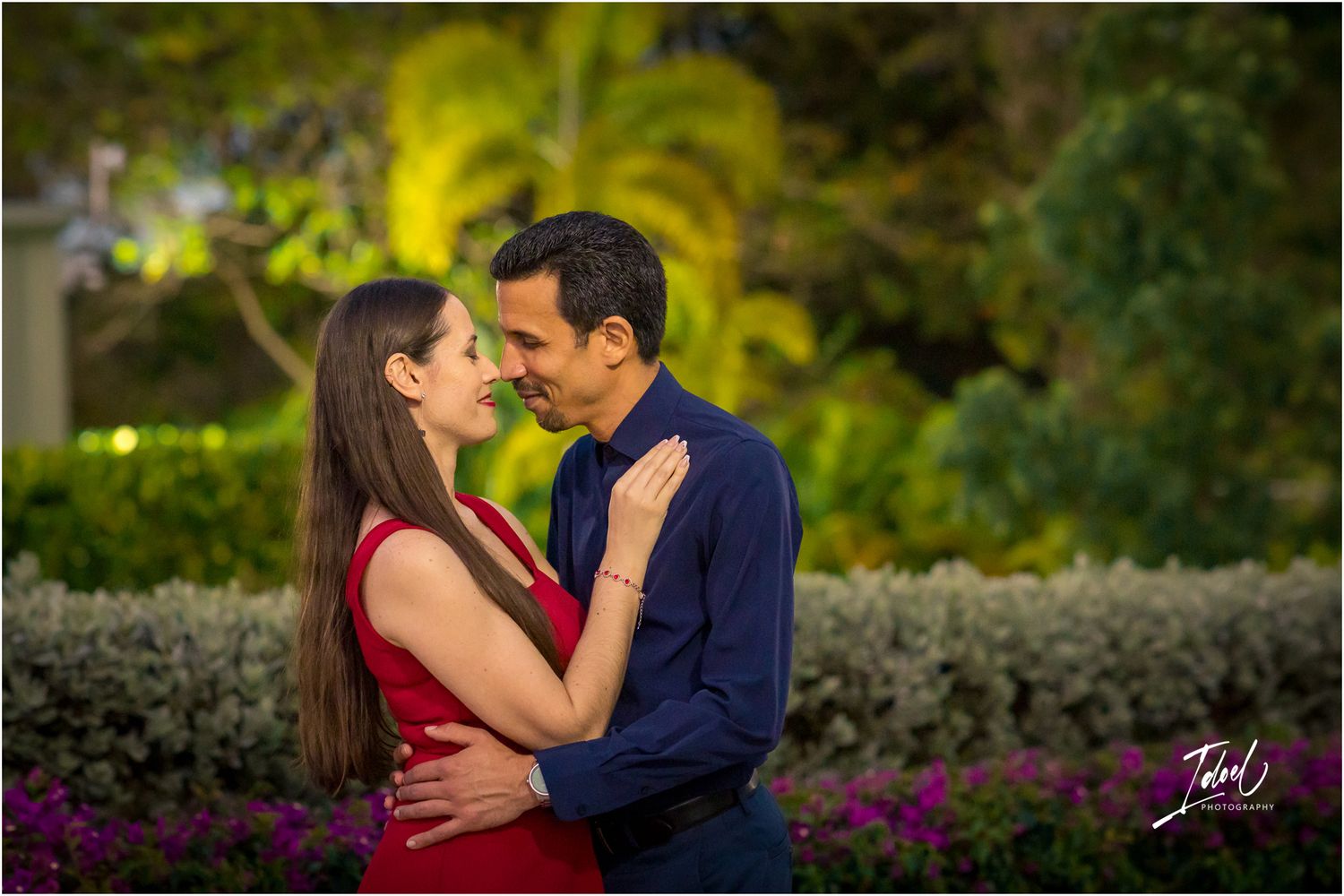 Romantic couple in red and blue embracing in a lush garden with colorful flowers at dusk.