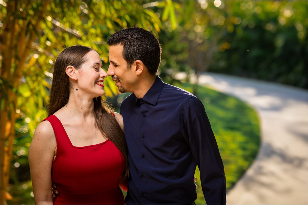 Couple smiling at each other outdoors, woman in red dress and man in navy shirt, surrounded by lush greenery.