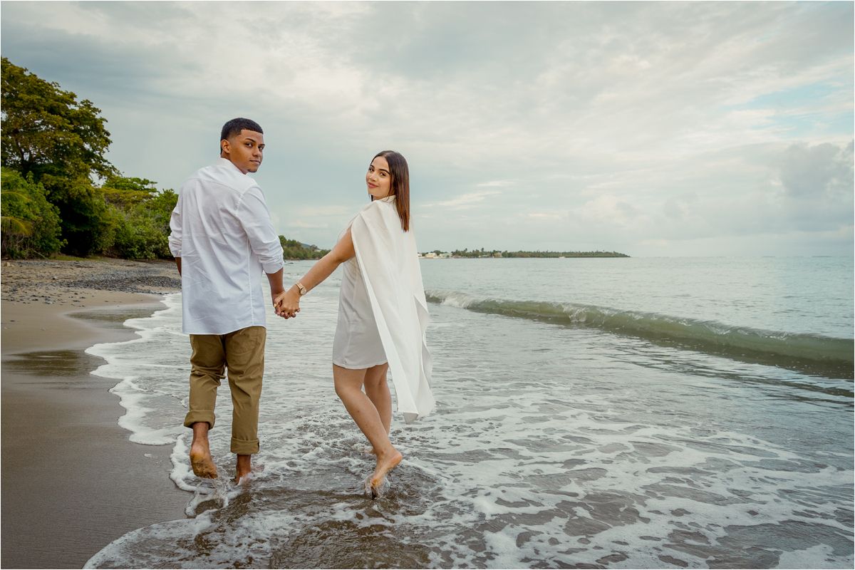 Couple holding hands walking barefoot on a tropical beach shore with gentle waves and lush green trees in background.