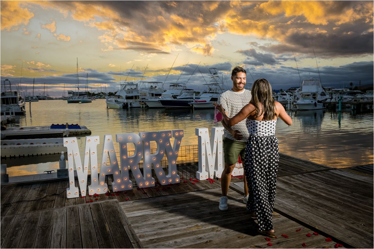 A couple stands on a marina dock at sunset with illuminated 