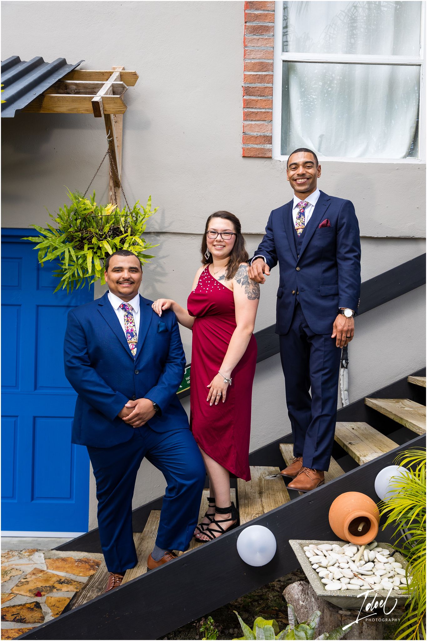 A group poses on outdoor stairs near a bright blue door wearing formal attire including navy suits and a burgundy dress.