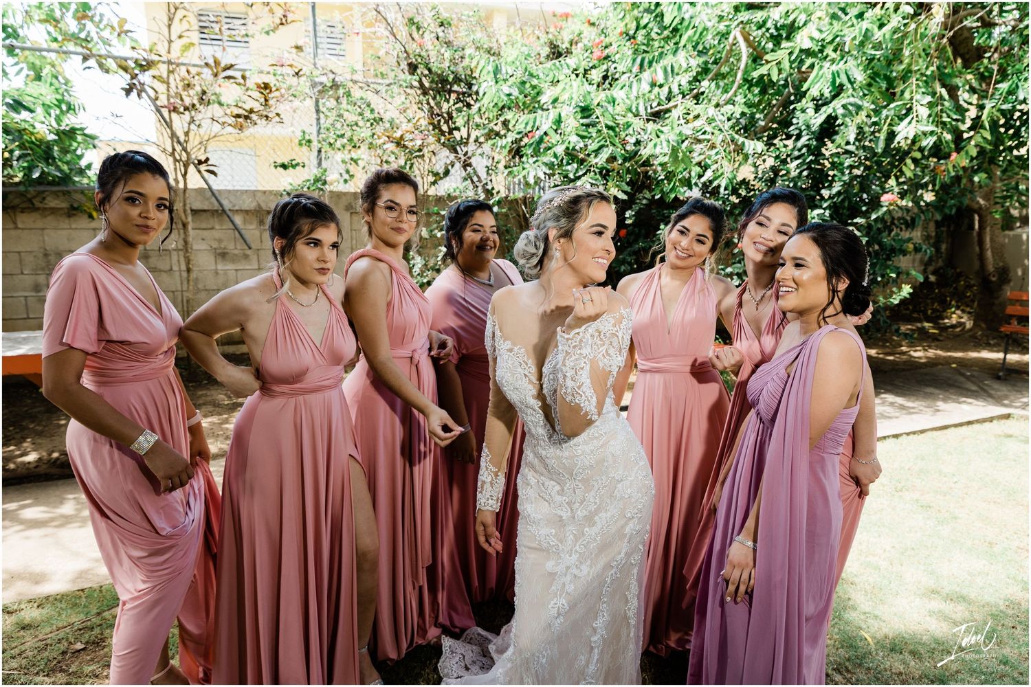 Bride in lace gown poses with bridesmaids wearing pink and mauve dresses in garden setting.
