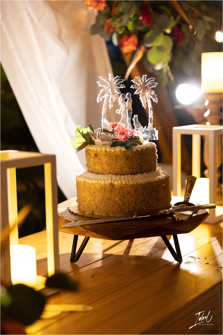 Two-tier wedding cake with palm tree toppers displayed on wooden stand with ambient lighting.