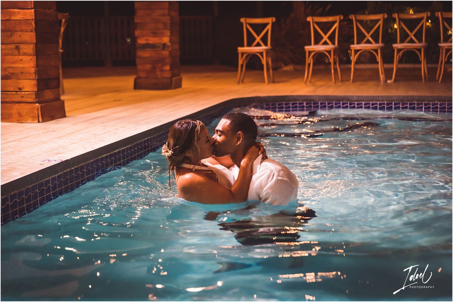 Romantic pool kiss scene at dusk with illuminated rustic decor and chairs in background.