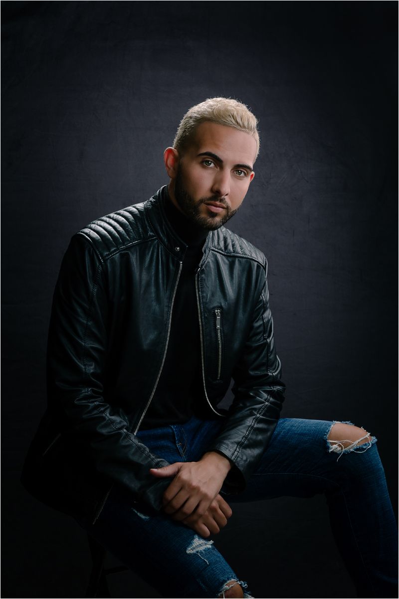 Dramatic studio portrait of someone in a black leather jacket and ripped jeans against a dark background.