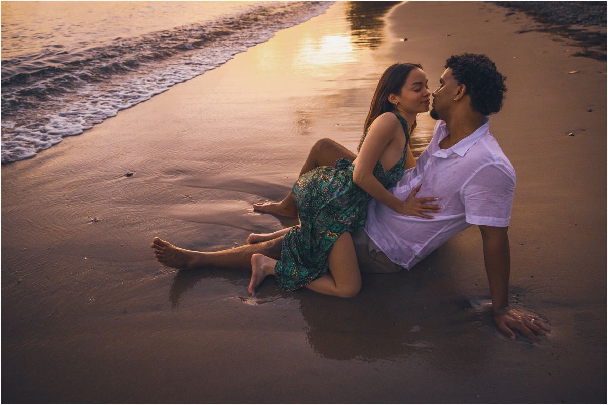 A romantic couple sits together on a sandy beach at sunset with waves lapping at the shore.